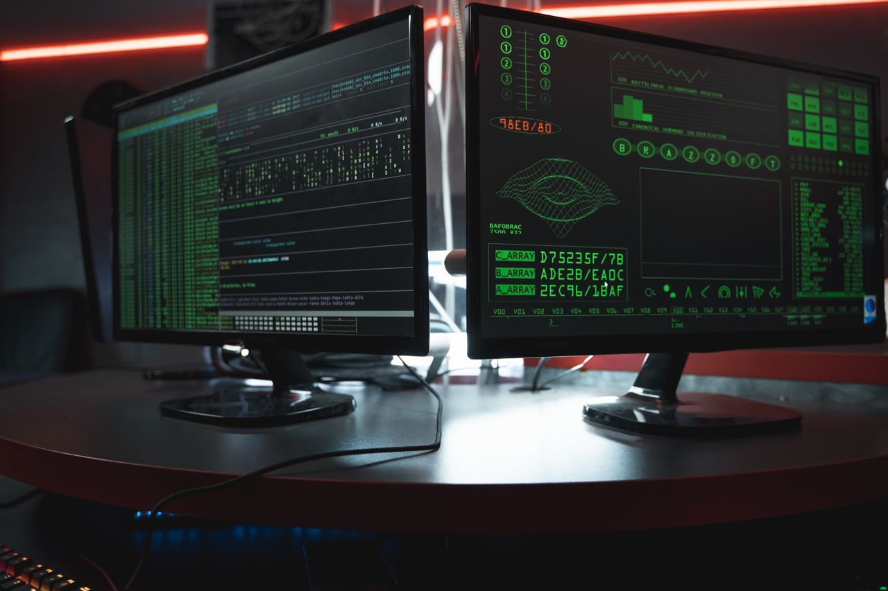 Close-up of dual computer monitors with green coding interfaces in a dark room, highlighting cyber security themes.